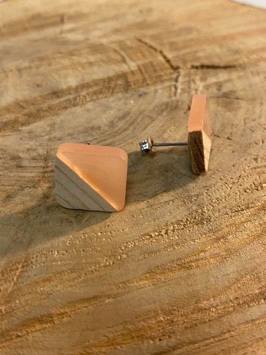 Ein Paar quadratische Ohrstecker aus Holz von wood stud mit einem diagonalen zweifarbigen Design aus hellem Naturholz und pfirsichfarbenem Harz, das auf einer Holzoberfläche präsentiert wird.