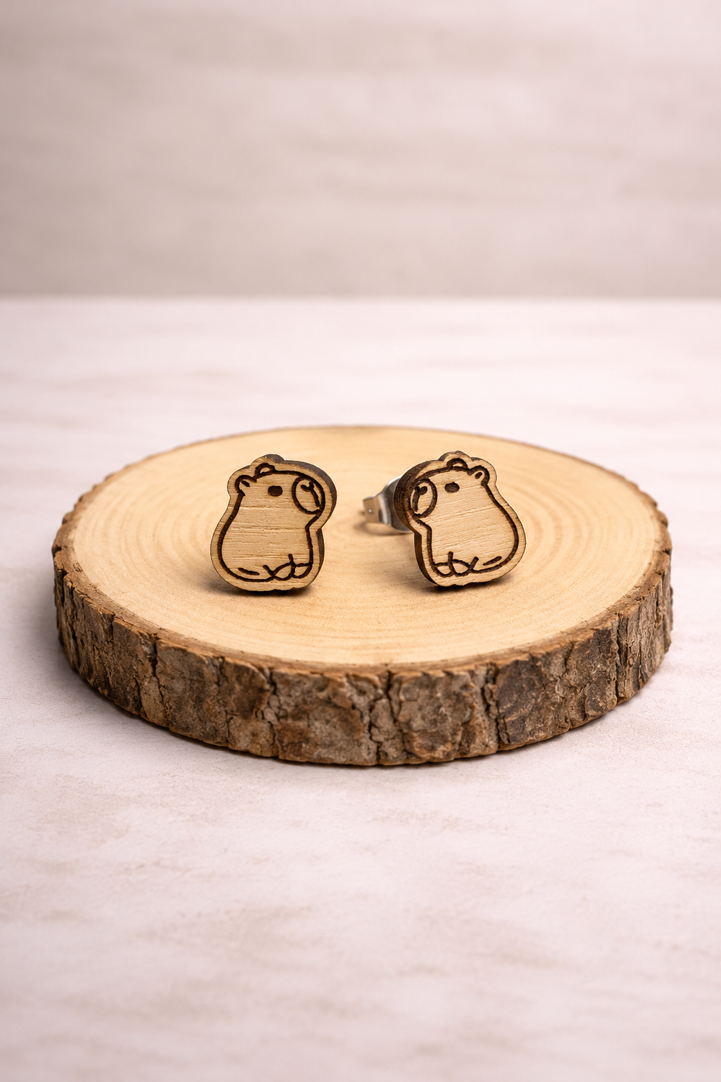 Capybara-Holzohrstecker: zwei sitzende Wasserschwein-Motive aus hellem Holz mit dunkler Gravur auf einer Baumscheibe.