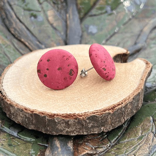 Ein Paar runder, rosafarben gesprenkelter Kork-Ohrstecker mit nickelfreien Edelstahlpfosten ruht auf einer glatten, runden Holzplatte vor einem sanft verschwommenen grünen und braunen Naturhintergrund.