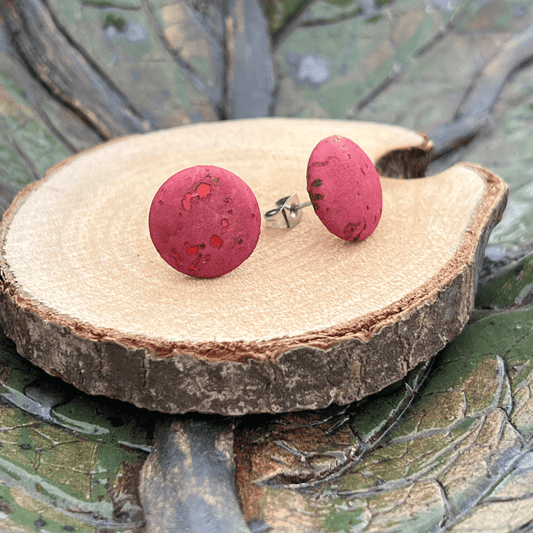 Ein Paar runder, roter Holznoppen-Ohrstecker mit dezenten dunklen Sprenkeln ruht auf einer glatten Holzscheibe vor grün-braunem Hintergrund - stilvoller und umweltfreundlicher Ohrschmuck.