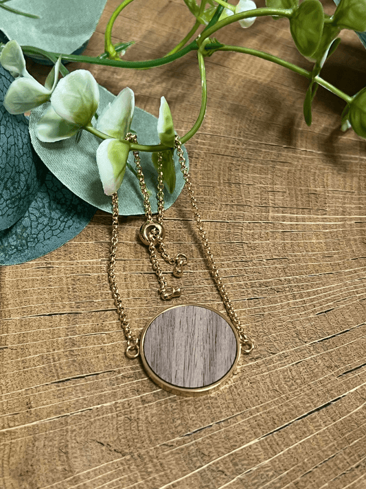 Eine goldene Halskette mit einem runden Holzanhänger, hypoallergenem Holzinlay und einem kleinen Giraffenanhänger wird auf Holz neben grünen Blättern und weißen Blumen präsentiert.