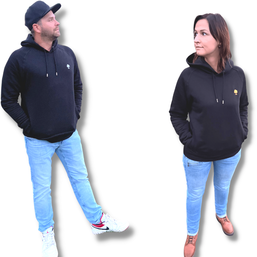 Zwei Personen stehen nebeneinander, beide tragen einen schwarzen Kapuzenpullover und hellblaue Jeans. Der Mann auf der linken Seite trägt eine schwarze Mütze und weiße Turnschuhe, die Hände in den Taschen. Die Frau auf der rechten Seite hat braunes Haar und trägt braune Schuhe. Beide schauen sich an.