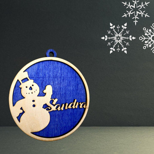 Ein Holzanhänger mit einer Schneemann-Silhouette und der ausgeschnittenen "Sandra" auf blauem Hintergrund. Schneeflocken akzentuieren die obere rechte Seite und machen diesen Baumschmuck zu einem herrlich festlichen Dekorationsstück.