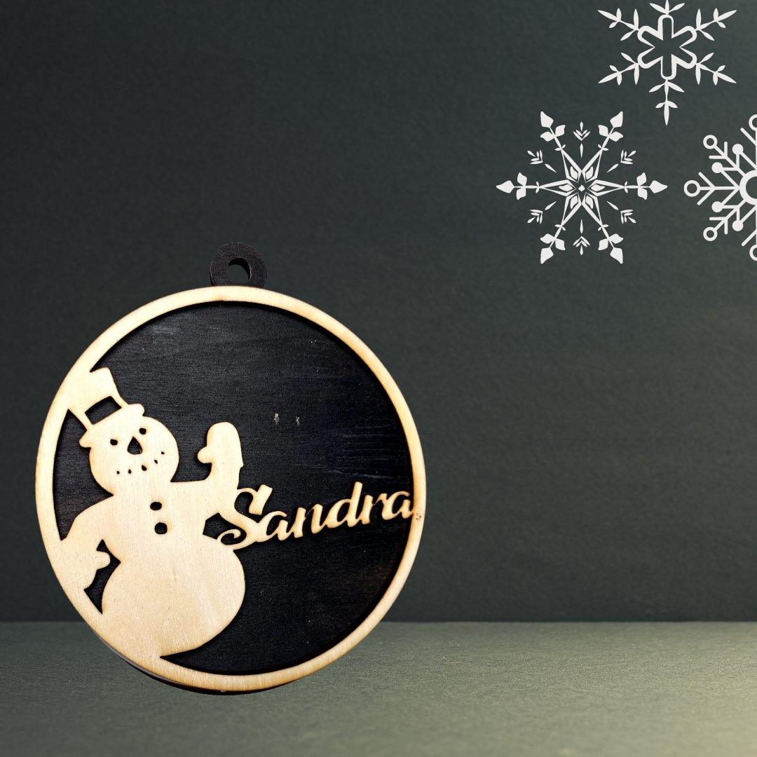 Ein runder Holzstollen Holzanhänger mit einem ausgeschnittenen Schneemann mit Hut und dem Namen Sandra, dargestellt auf einem dunklen Hintergrund mit weißen Schneeflocken - ideal als personalisierte 3D-Weihnachtskugel Baumschmuck.