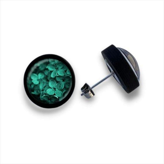 Ein Paar runde Ohrstecker aus Holz von wood stud, jeder mit einer schwarzen Umrandung und kleinen grünen Blumen unter einer Cabochon-Glaslinse. Ein Ohrring zeigt das Design, während der andere seine Birkenholzseite zeigt.