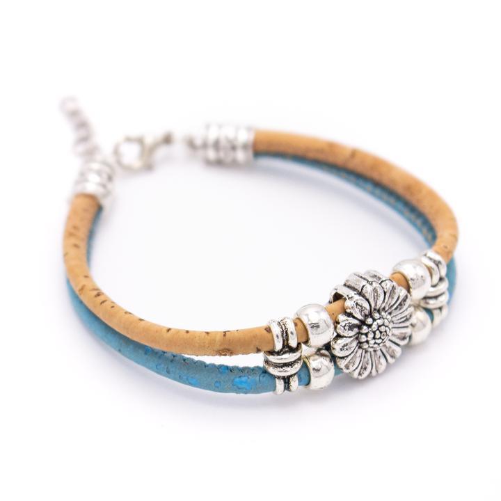Das Holzarmband besteht aus einem hellbraunen Korkstrang und einem blauen Strang, der mit nickelfreien Silberperlen aus Edelstahl und einem silbernen Blumenanhänger in der Mitte verziert ist. Abgebildet auf weißem Hintergrund - ideal für Fans von umweltfreundlichen Korkarmbändern.
