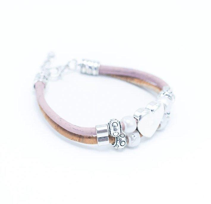 Ein veganes Lederarmband mit Holznieten in Rosa und Hellbraun, mit silbernen und perlenartigen Perlen, mit einem weißen herzförmigen Anhänger in der Mitte, auf weißem Hintergrund.