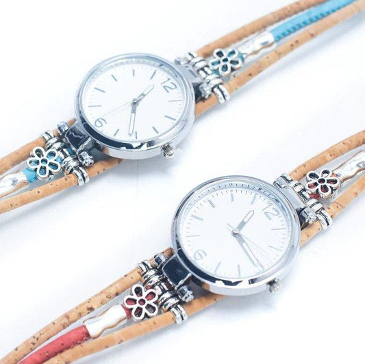 Zwei Armbanduhren mit Holzstiften, runden weißen Zifferblättern und silbernen Rahmen und braunen Korkarmbändern, die mit silbernen Blumenanhängern und farbigen Akzenten verziert sind, heben nachhaltige Materialien und natürliches Design hervor und werden nebeneinander auf einem weißen Hintergrund präsentiert.