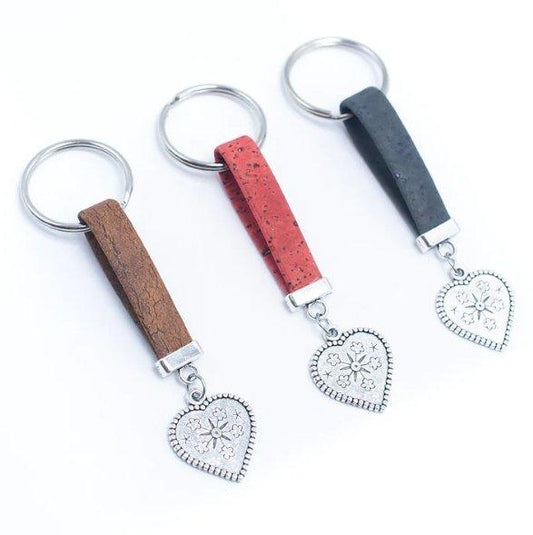 Drei umweltfreundliche Kork-Schlüsselanhänger von Holzständer, jeweils mit einem Lederband in Braun, Rot und Schwarz und silbernem herzförmigen Anhänger mit Blumenmuster, liegen nebeneinander auf weißem Hintergrund für extra Nachhaltigkeit.