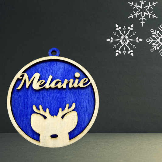 Ein runder, blauer Holzanhänger von wood stud mit einer hölzernen Umrandung, auf der "Melanie" und ein Rentierkopf abgebildet sind. Weiße Schneeflocken akzentuieren die obere rechte Ecke. Diese 3D-Weihnachtskugel ist personalisierbar.