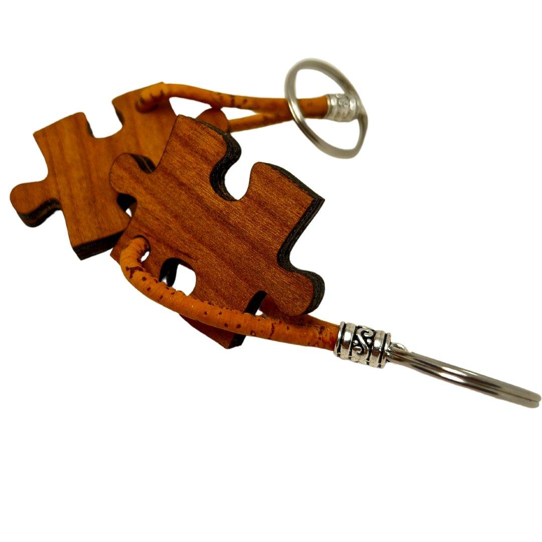 Zwei Holz-Schlüsselanhänger in Puzzleform von Holzstollen, verbunden durch tan Lederbänder, zeigen natürliche Maserung und nachhaltiges Design. Jeder ist an einem silbernen Metallring befestigt.