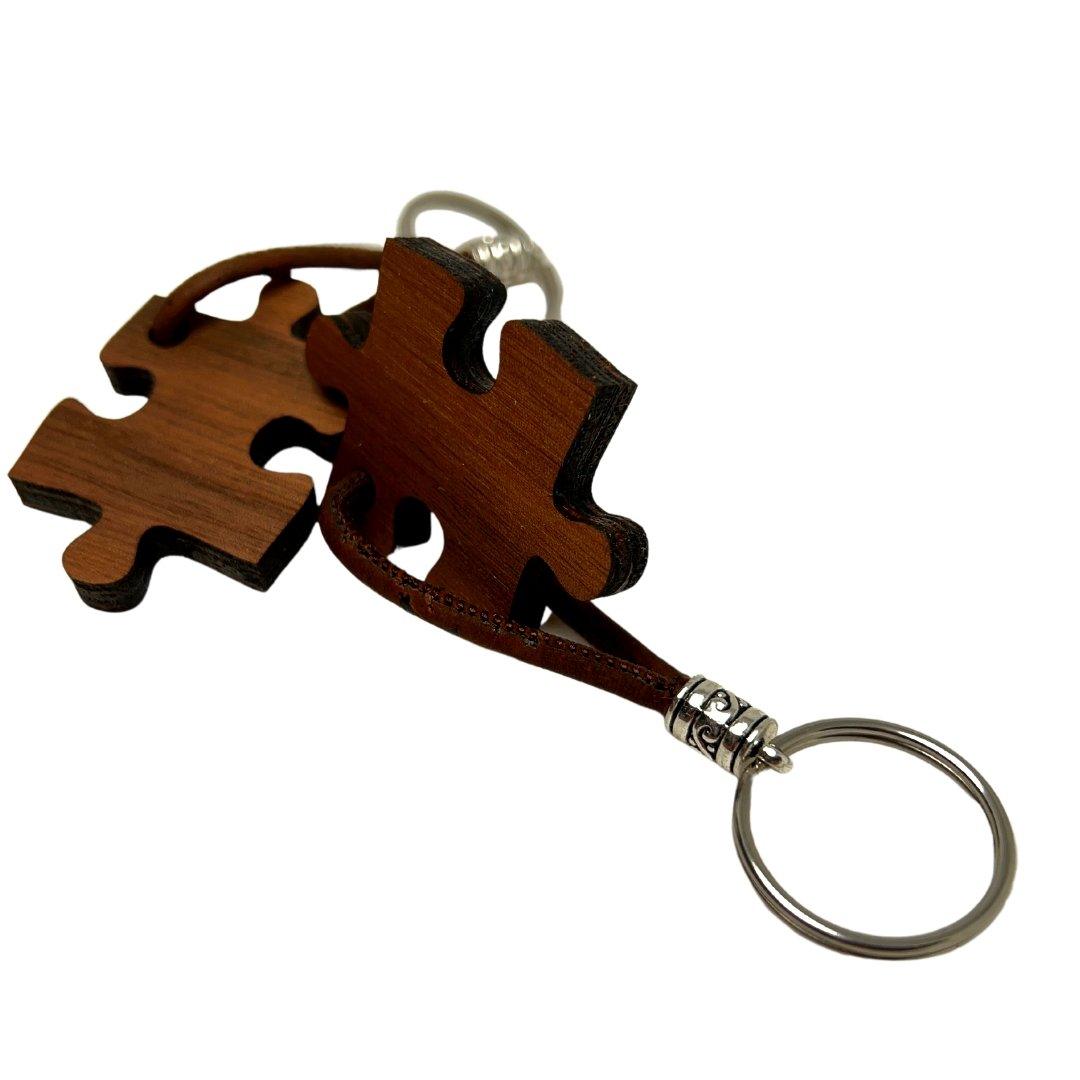Zwei Holz-Schlüsselanhänger von wood stud in Puzzleteilform sind mit einem braunen Lederband verbunden. Die natürliche Maserung macht jedes Stück einzigartig, silberne Metallringe halten Ihre Schlüssel stilvoll und sicher.