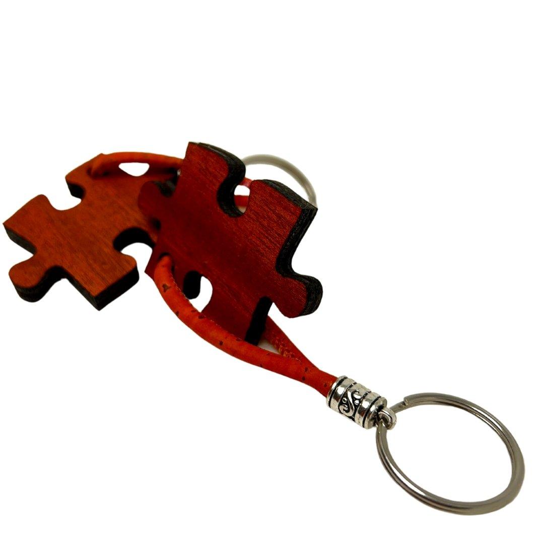 Ein Holz-Schlüsselanhänger von wood stud mit zwei ineinandergreifenden Puzzlestücken, deren natürliche Maserung durch nachhaltiges Design, rote Lederstreifen und silbernen Schlüsselring stilvoll hervorgehoben wird.