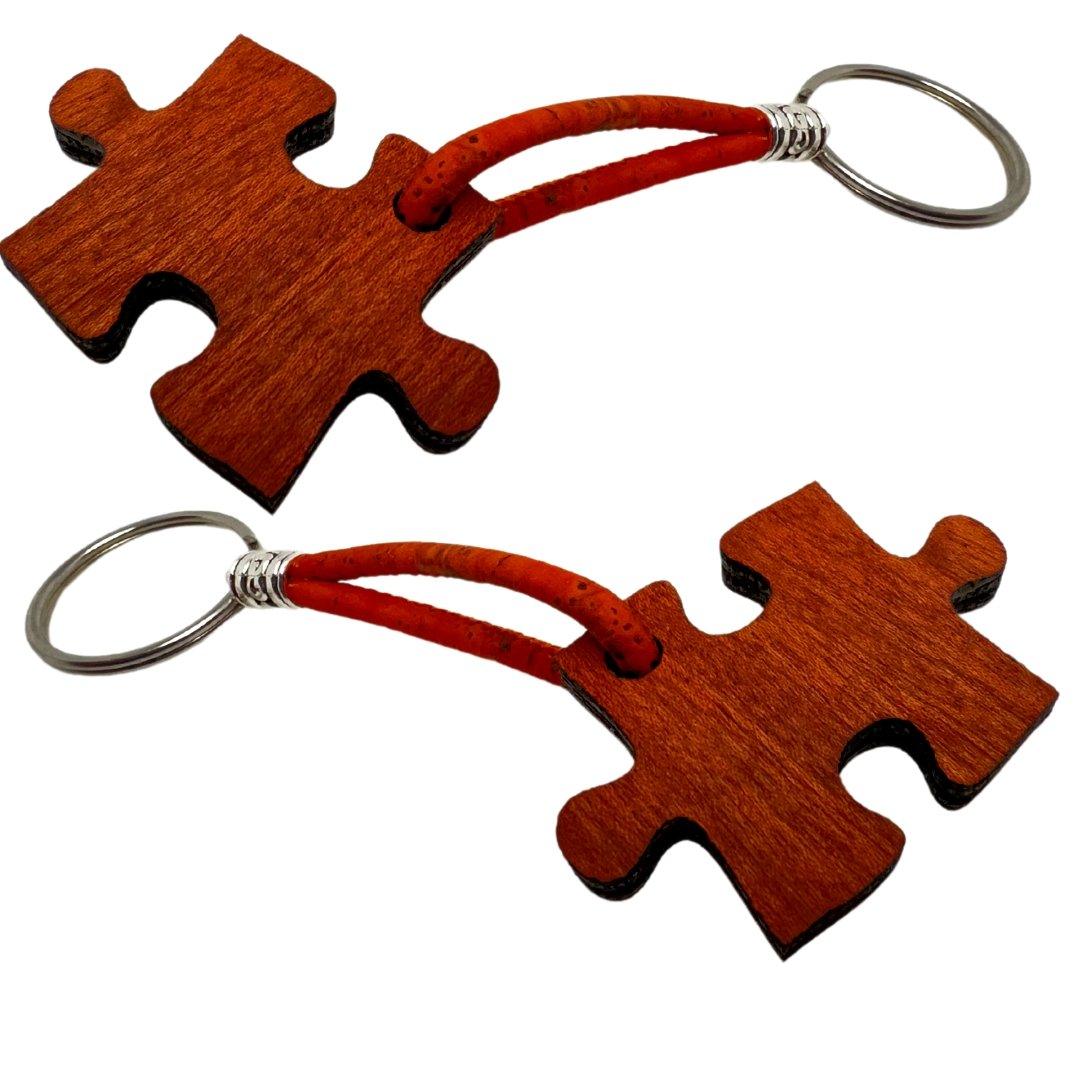 Zwei Holzstollen Holz-Schlüsselanhänger in Puzzleform mit natürlicher Maserung, roten Lederloops und silbernen Ringen liegen nebeneinander auf weißem Hintergrund.