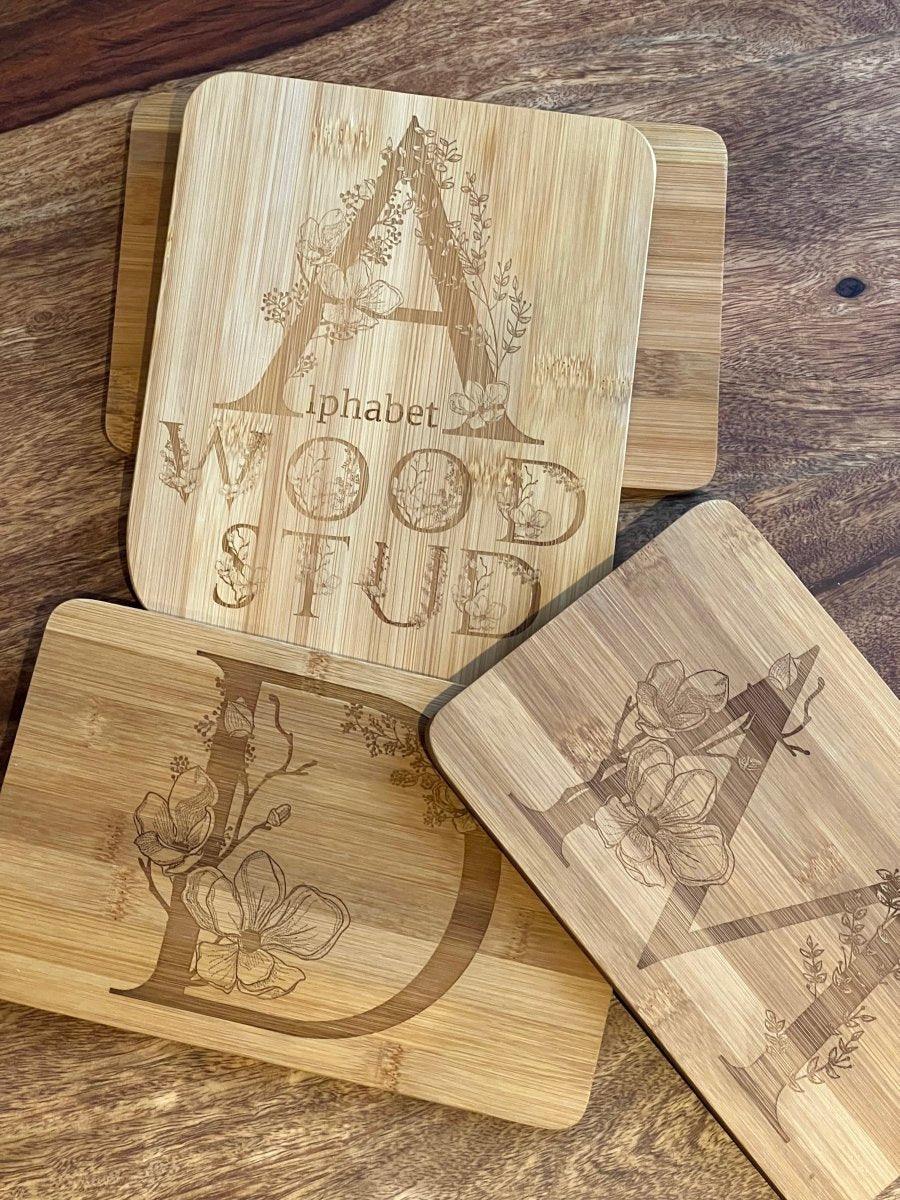 Vier Holzstollenplatten zeigen die eingravierten Blumenbuchstaben A, D und V sowie den Schriftzug "Alphabet Wood Stud" auf einer Holzoberfläche. Jedes Design weist eine detaillierte botanische Gravur aus Holzstiften auf - eine charmante Geschenkidee.
