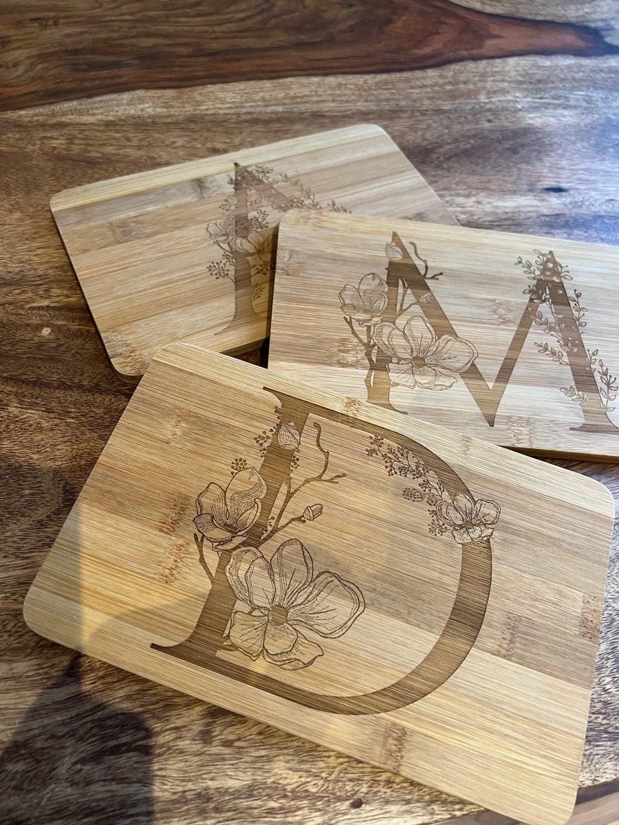 Drei rechteckige Holzbretter mit Holzstollen, jedes mit einem großen D, M oder A und detaillierten floralen Holzgravur-Motiven graviert, sind auf Holz geschichtet - eine einzigartige Geschenkidee.