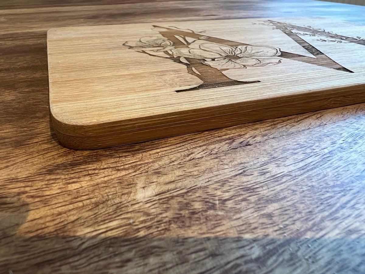 Ein Schneidebrett aus Holz mit einem Blumenmuster und dem Buchstaben "A" ruht auf einer strukturierten Holzoberfläche, was es zu einer charmanten Geschenkidee macht.