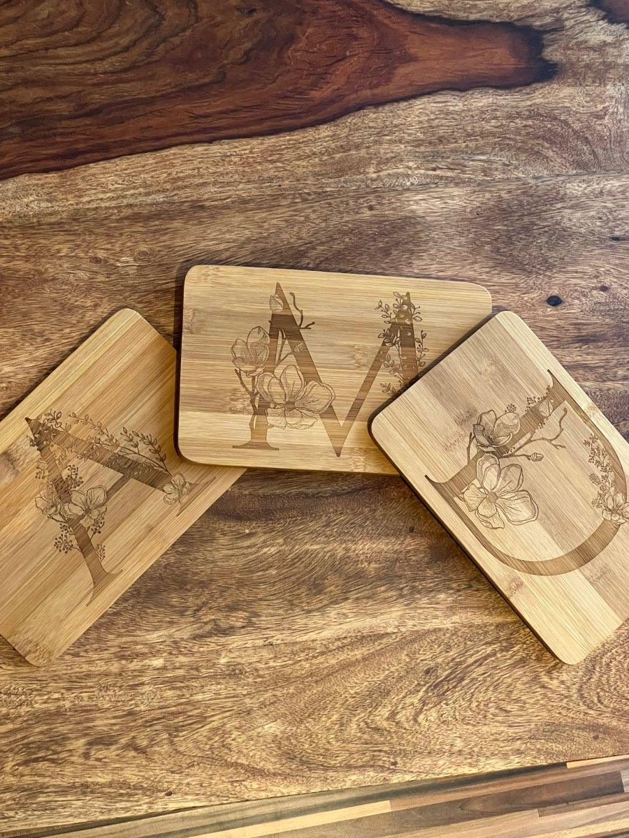 Drei Schneidebretter mit Holzstiften zeigen exquisite Blumenmotive aus Holzgravur mit den auffälligen Buchstaben A, M und C. Jedes Brett zeigt ein einzigartiges Blumenmotiv, das mit dem jeweiligen Buchstaben verwoben ist - eine charmante Geschenkidee für jeden Anlass.