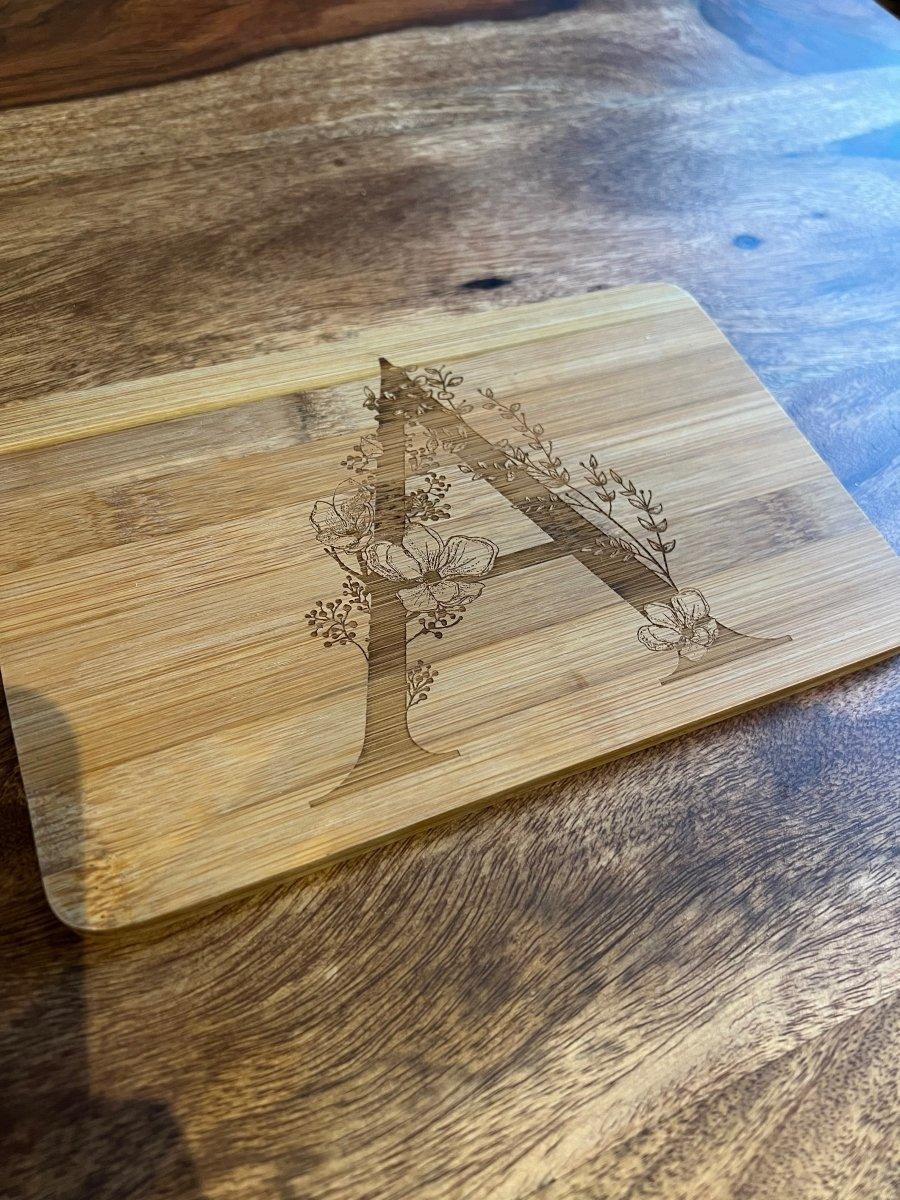 Ein hölzernes Schneidebrett mit einem großen, eingravierten Buchstaben A mit floralen und botanischen Motiven ruht auf einer hölzernen Oberfläche - eine einzigartige Holzgravur und perfekte Geschenkidee.