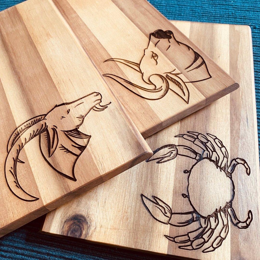 Drei gravierte Schneidebretter aus hochwertigem Holz von wood stud mit detailreicher Sternzeichen-Gravur (Steinbock, Stier, Krebs) kunstvoll auf tealfarbenem Stoff präsentiert.