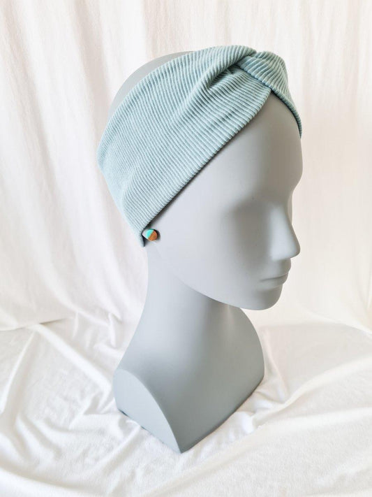 Eine graue Schaufensterpuppe trägt ein hellblaues, geripptes Twister-Stirnband aus Holz mit einer Verdrehung am oberen Ende vor einem weiß drapierten Hintergrund.