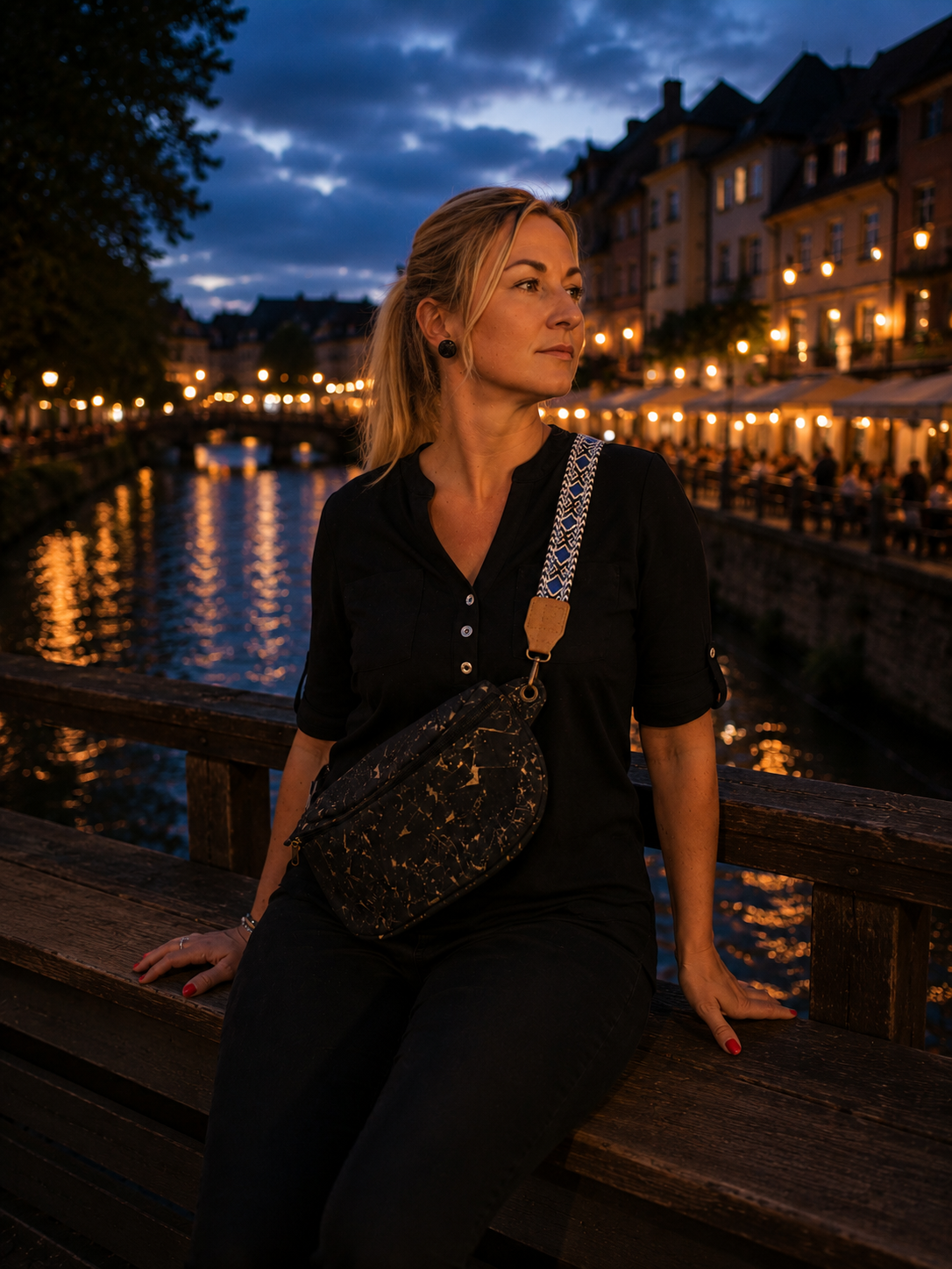 Eine Frau sitzt in der Abenddämmerung auf einer Holzbrüstung am Ufer eines Kanals, beleuchtet von Lichterketten, die von den Gebäuden am Ufer herabstrahlen. Sie trägt ein schwarzes Outfit und eine stilvolle Bauchtasche mit Holznieten in Schwarz und Gold, die sie quer über den Körper trägt, und blickt nachdenklich zur Seite.
