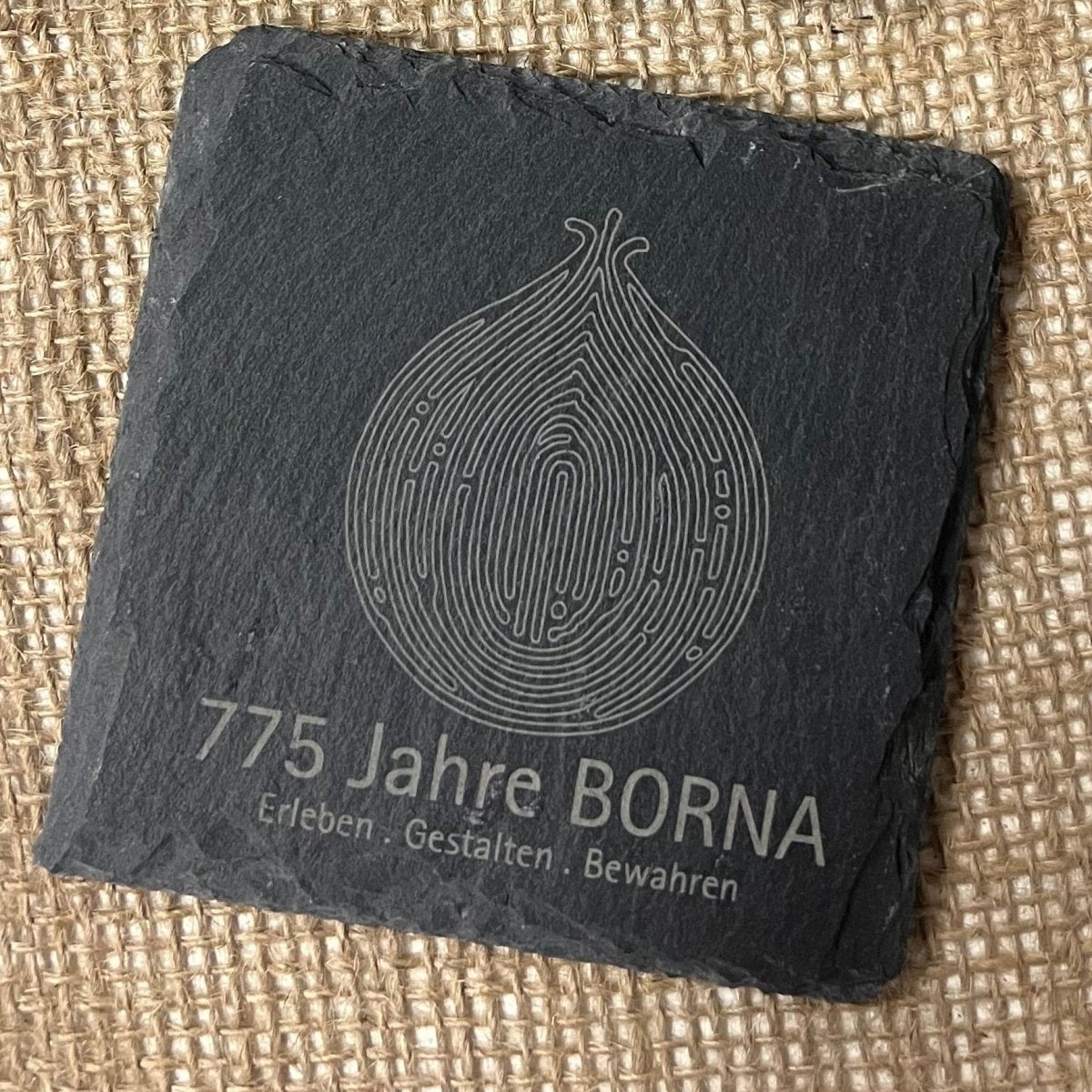 Quadratischer schwarzer Schieferuntersetzer Borna aus Holzstollen auf Sackleinen, mit einer stilisierten Zwiebel und eingraviertem Text "775 Jahre BORNA. Erleben - Gestalten - Bewahren." Dieses in Thüringen handgefertigte Stück ist Teil der Jubiläumskollektion.